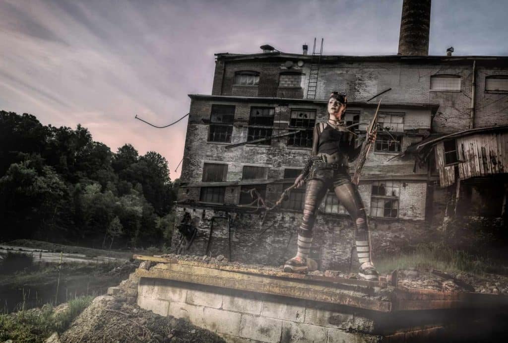 Cosplay model in urban decay setting, holding a weapon, showcasing dramatic lighting and industrial background, representing creative photography style by Nimble Dragon Studios.