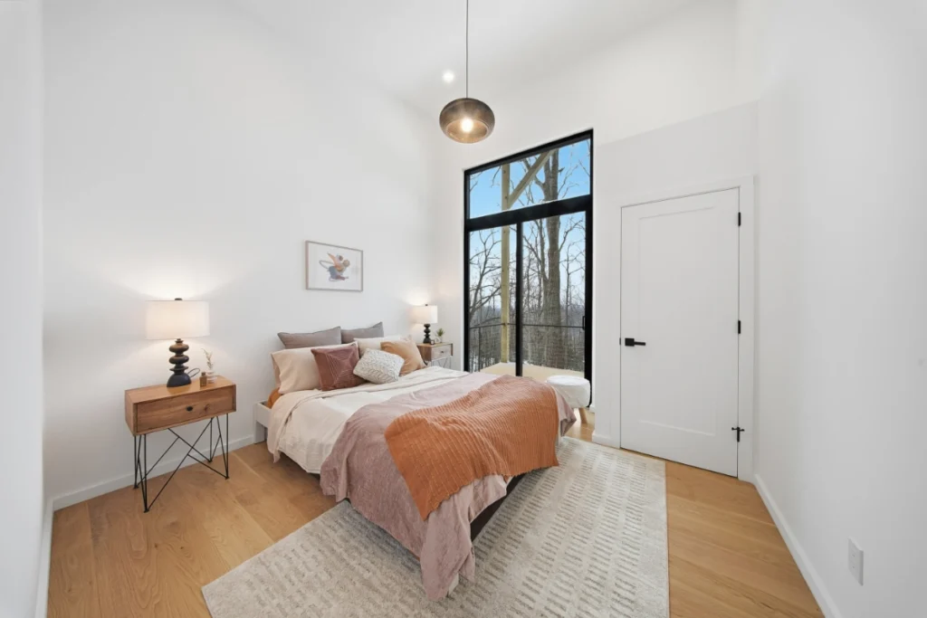 Modern bedroom with wooden floors, bed with orange throw, nightstand lamp, and large black-framed window.
