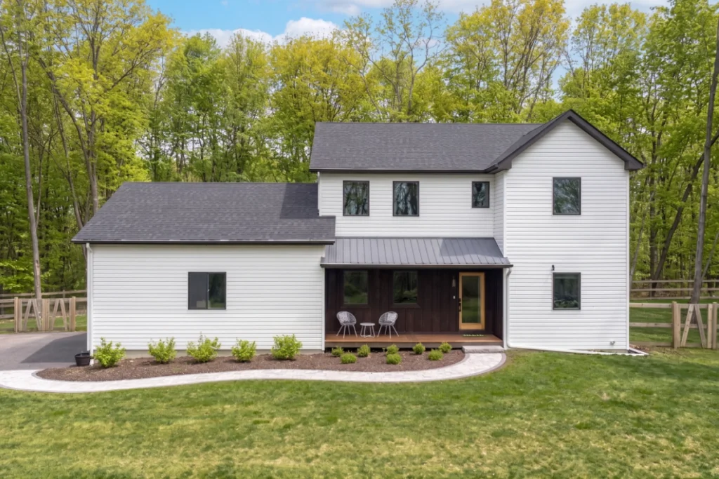 Modern white farmhouse with covered porch and landscaping in wooded setting.