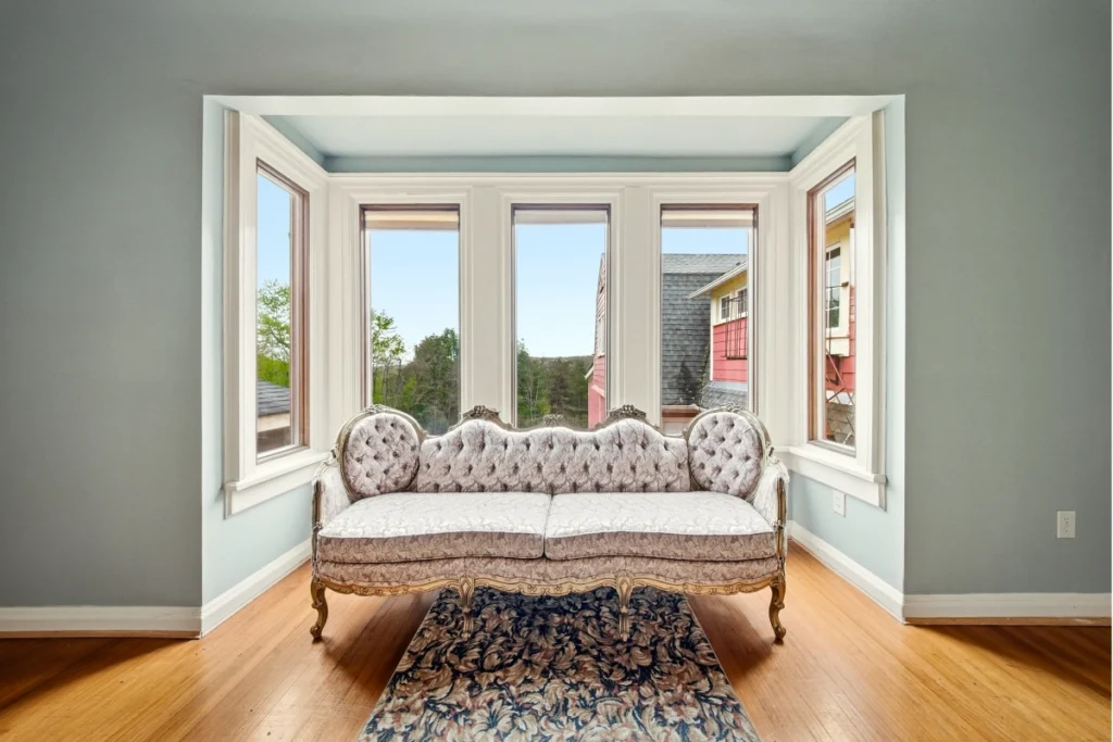Ornate settee positioned in bay window overlooking scenic landscape and neighboring homes.