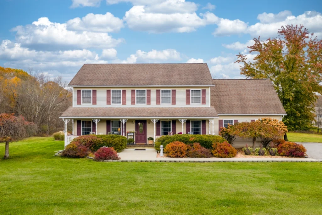 Classic two-story farmhouse with red shutters and fall landscaping on green lawn.