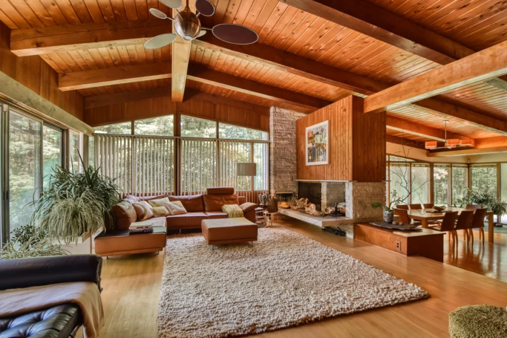 Warm mid-century modern living room with wood ceilings and fireplace overlooking forest.